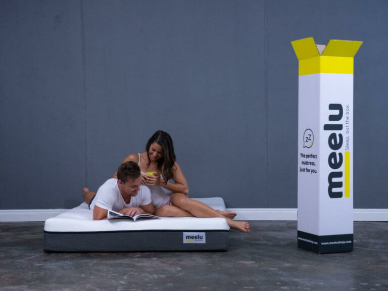 an image of a woman holding a coffee cup sitting next to a man lying on his stomach and reading a book on top of the Meelu mattress, pictured next to the box that the mattress comes in