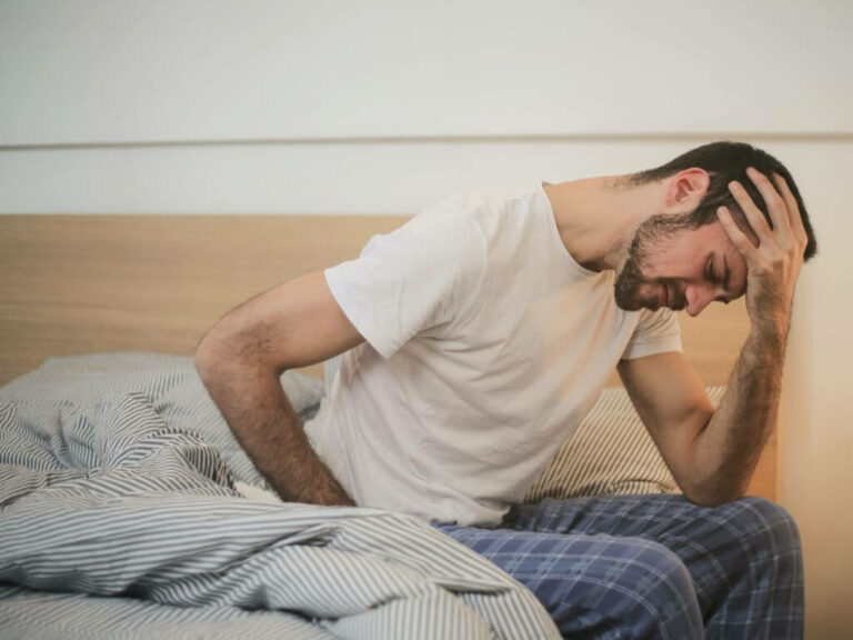 A man sitting on the side of his unmade bed, in his pyjamas, appearing to be experiencing back pain, and holding his head.